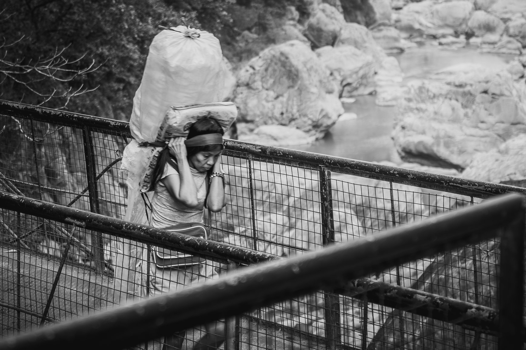 A woman carries a large load of goods on her back across a bridge.