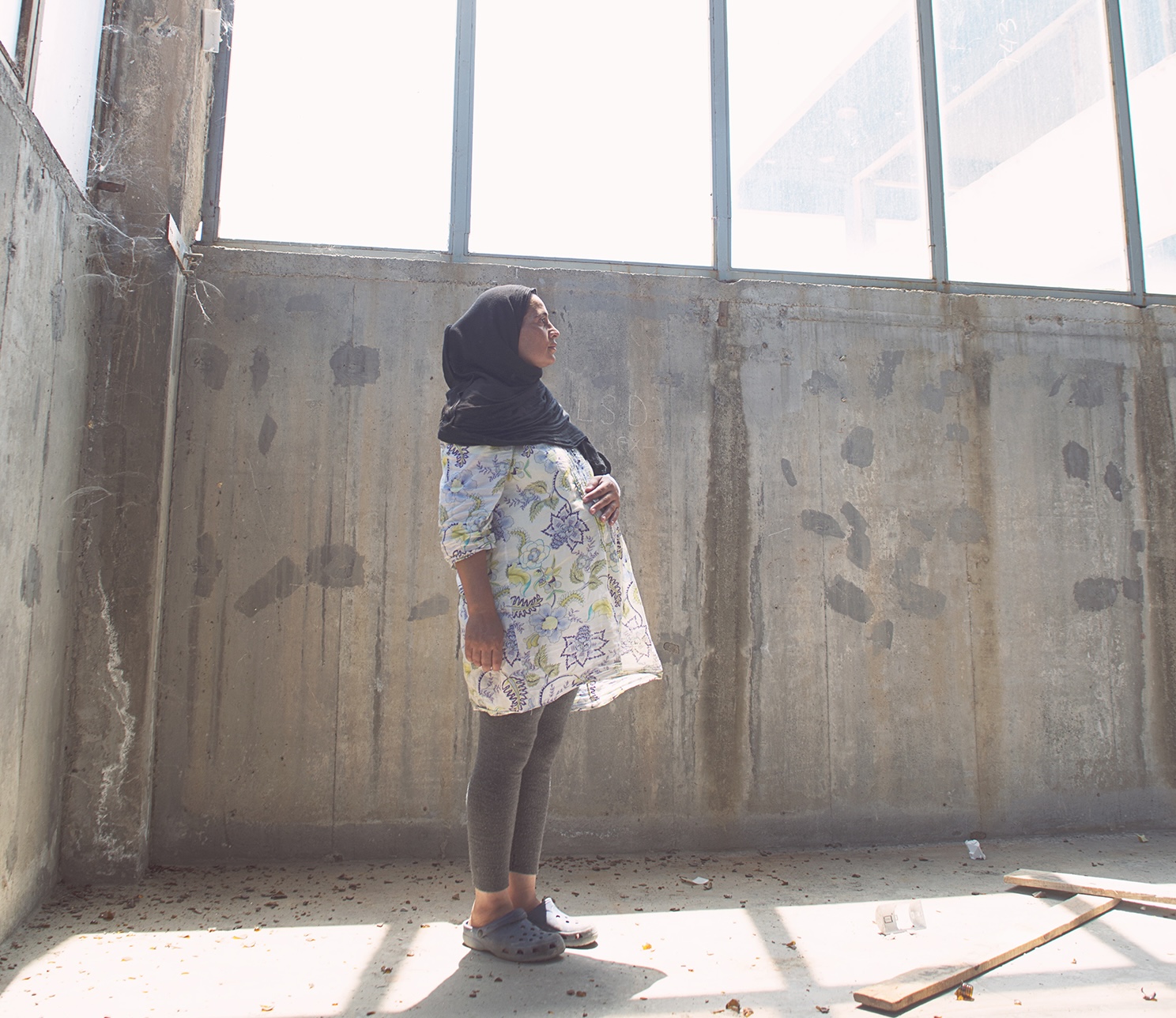 Pregnant woman in a headscarf stands in a barren concrete room with the sun streaming in.